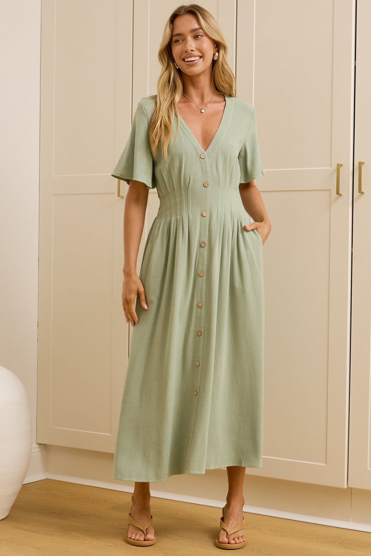 A woman stands in a bright room wearing a light sage green midi dress with short sleeves, a V-neckline, and front buttons. A large mirror on the left reflects her side profile, and cream-colored cabinets are in the background.