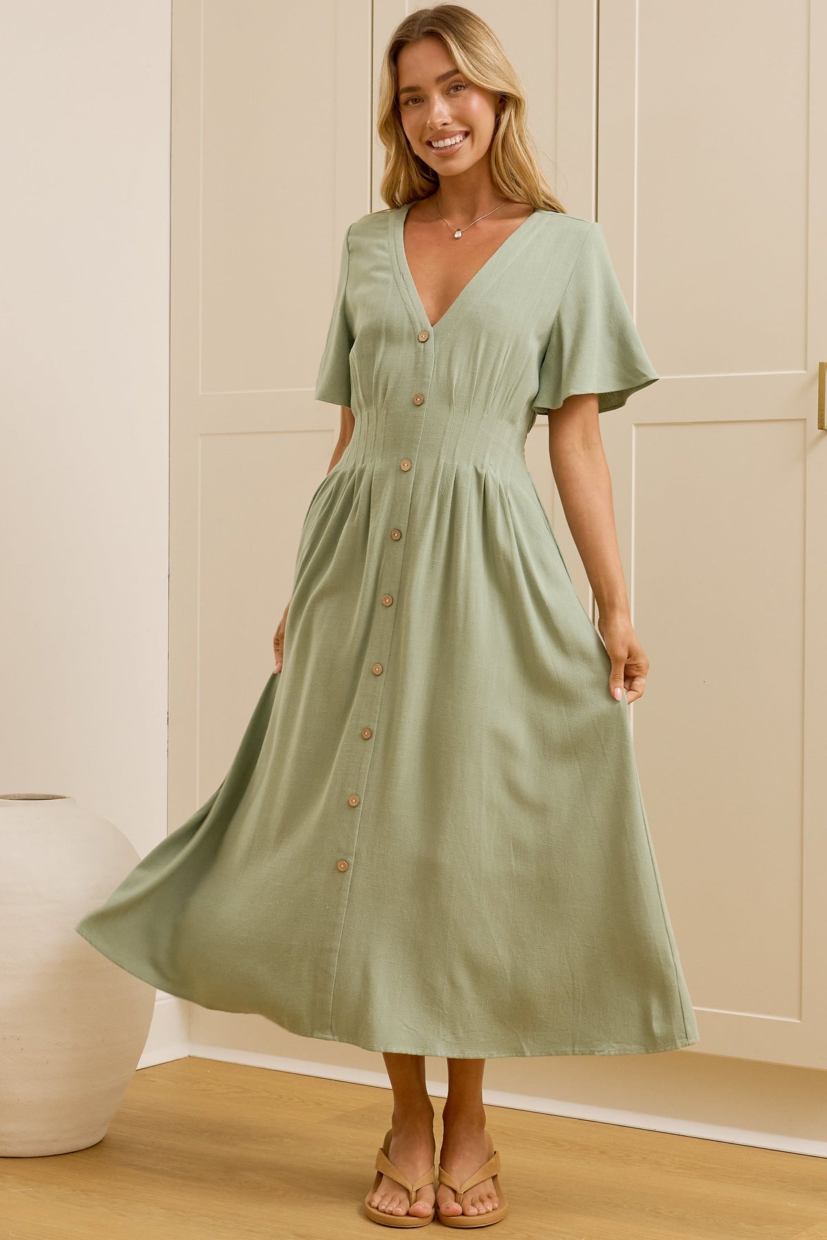 A woman modeling a light sage green midi dress with short flutter sleeves, a V-neckline, and button details down the front, standing in a room with cream-colored wooden wardrobes and a mirror showing her side profile.