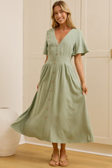 A woman modeling a light sage green midi dress with short flutter sleeves, a V-neckline, and button details down the front, standing in a room with cream-colored wooden wardrobes and a mirror showing her side profile.