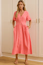 A woman wearing a coral pink midi dress with short flutter sleeves and a button-front detail stands in a bright room with white wardrobes, with her reflection visible in a large mirror on the left.