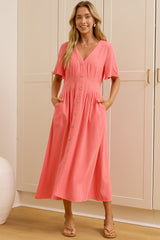 A woman wearing a coral pink midi dress with short sleeves and a V-neck stands in a bright room with white paneled walls and a wooden floor. A large mirror on the left reflects her side profile, and two white decorative vases sit on the floor.