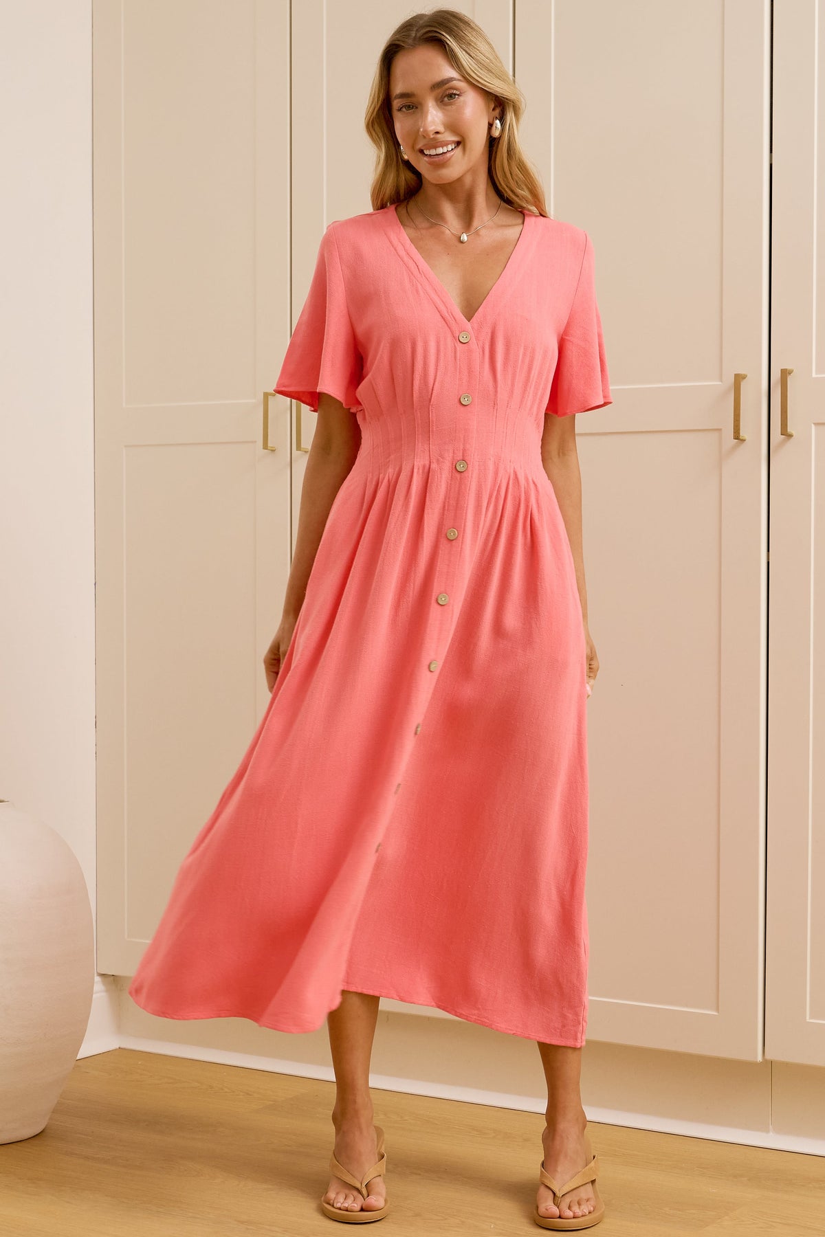 A woman modeling a coral pink midi dress with short sleeves and a V-neckline, standing in a bright room with white wardrobes and a large mirror showing her reflection.