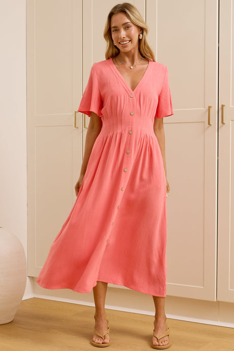 A woman modeling a coral pink midi dress with short sleeves and a V-neckline, standing in a bright room with white wardrobes and a large mirror showing her reflection.