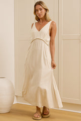 A woman stands in a bright room wearing a sleeveless cream maxi dress with a V-neck and gathered waist. A large mirror on the left reflects her side profile, and two white ceramic vases sit on the floor against cream paneled walls.