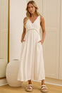 A woman wearing a sleeveless white maxi dress with a V-neck and side pockets stands in a bright room with cream-colored cabinets and a large mirror showing her reflection.