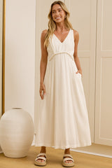 A woman wearing a sleeveless white maxi dress with a V-neckline and side pockets stands in a bright room with cream-colored cabinets, a large gold-framed mirror showing her reflection, and a white ceramic vase on a light wood floor.