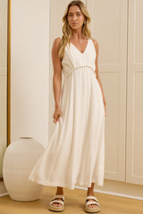 A woman wearing a sleeveless white maxi dress with a V-neckline and tie waist stands in a bright room with cream-colored wardrobes and a large gold-framed mirror showing her side profile.