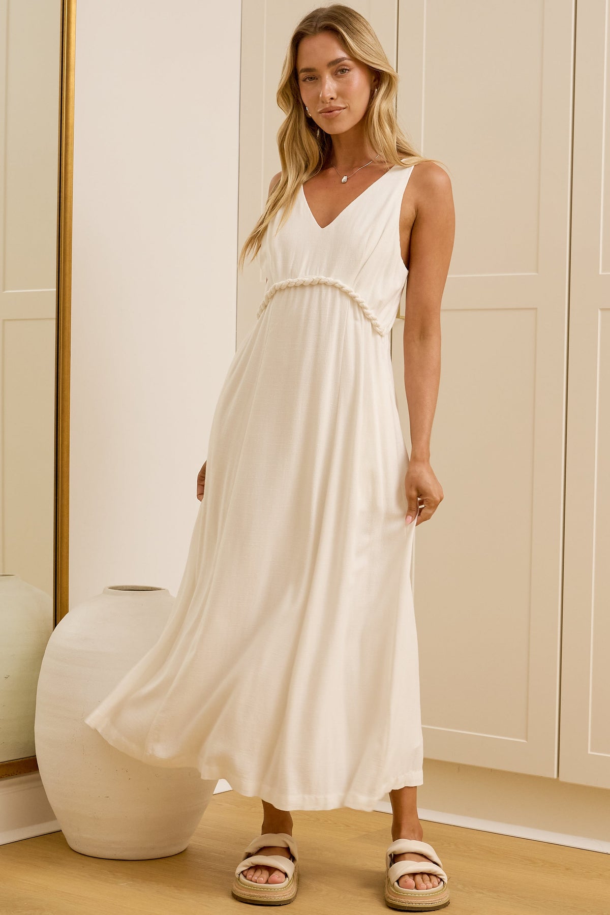 A woman wearing a sleeveless white maxi dress with a V-neckline and side pockets, standing in a bright room with cream-colored cabinets and a large gold-framed mirror showing her reflection. 