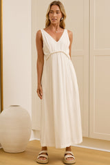 A woman wearing a sleeveless white maxi dress with a V-neckline and gathered waist stands in a bright room with cream-colored cabinets, a large mirror showing her side profile, and a white vase on the floor.