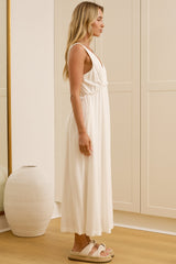 A woman stands in profile wearing a sleeveless white maxi dress and light sandals in a bright room with cream built-in wardrobes, a large gold-framed mirror showing her reflection, and two white vases on a light wood floor.