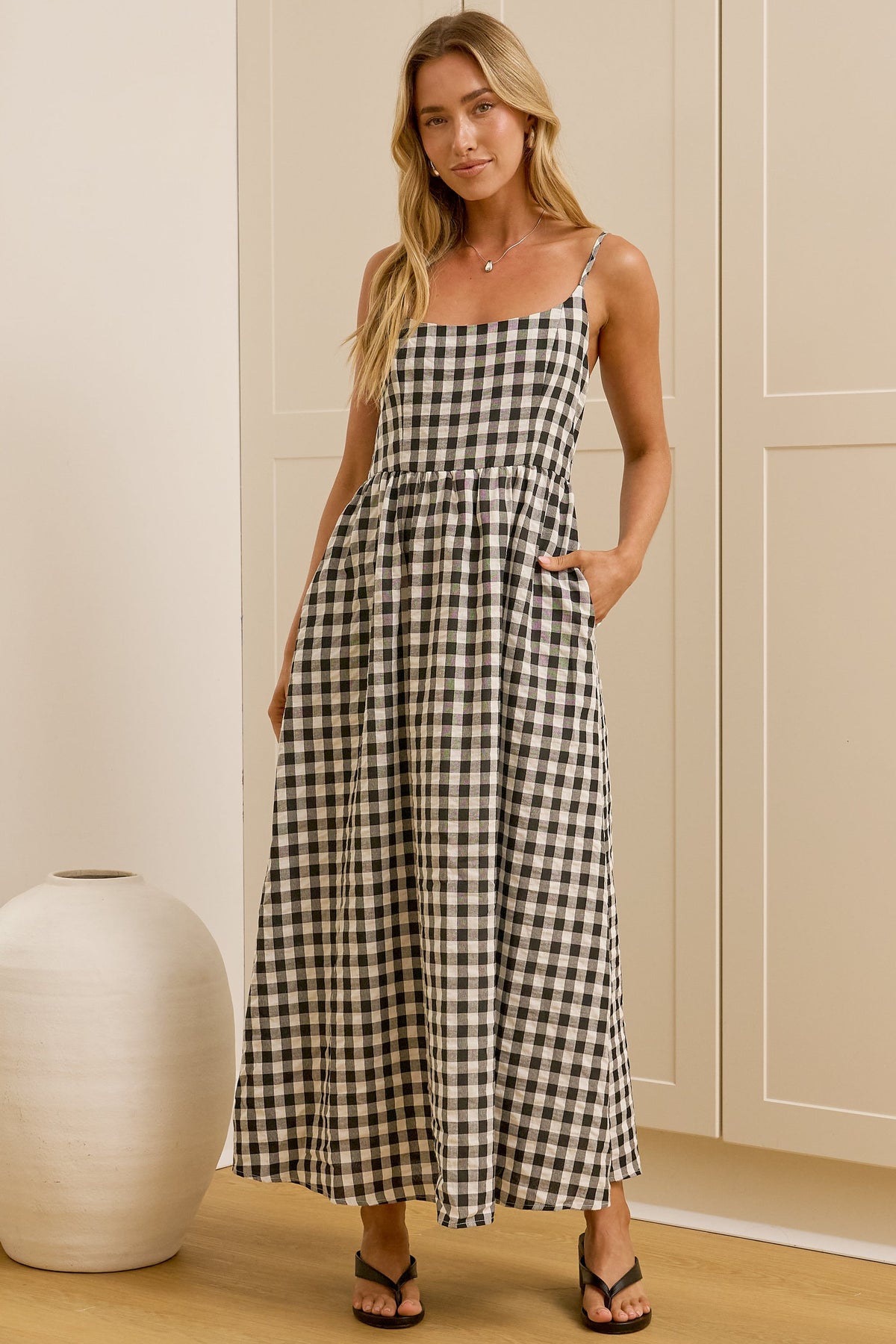 A woman modeling a black and white gingham maxi dress with thin spaghetti straps, a square neckline, and side pockets, standing in a brightly lit room with a white ceramic vase on the left.