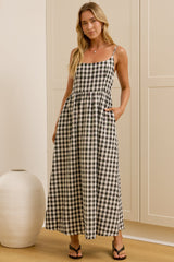 A woman modeling a black and white gingham maxi dress with thin spaghetti straps, a square neckline, and side pockets, standing in a brightly lit room with a white ceramic vase on the left.