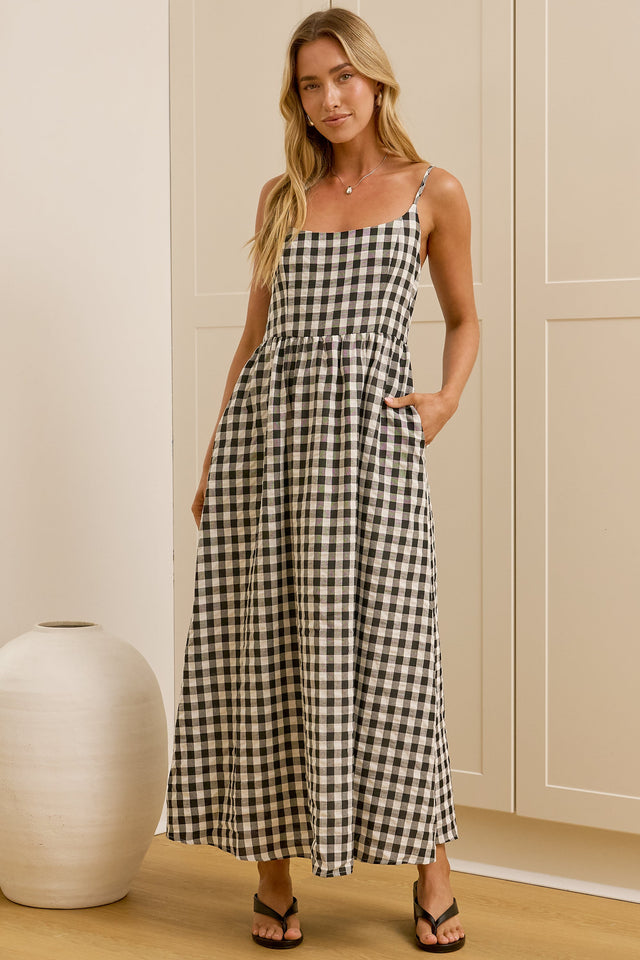 A woman modeling a black and white gingham maxi dress with thin spaghetti straps, a square neckline, and side pockets, standing in a brightly lit room with a white ceramic vase on the left.