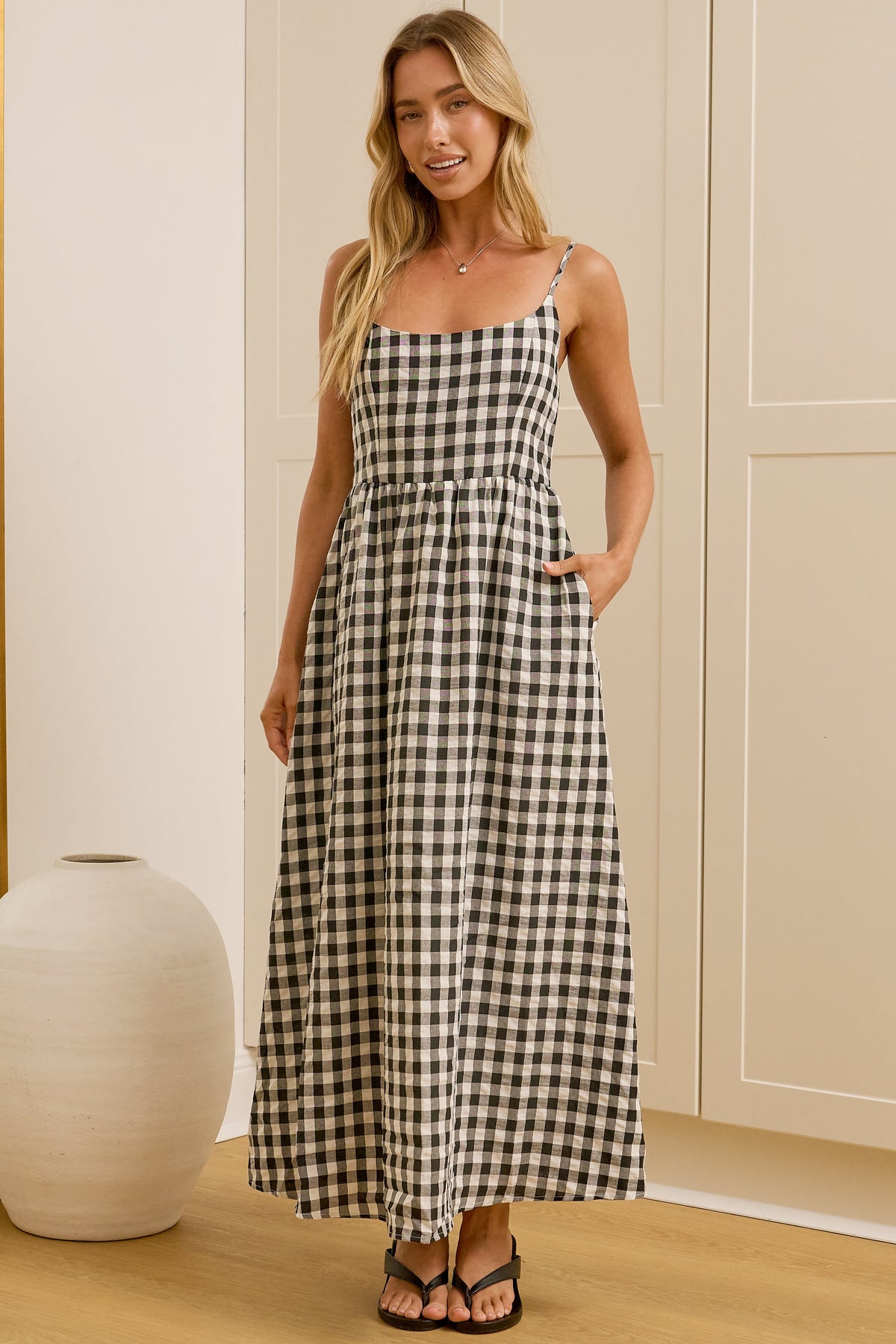 A woman modeling a black and white gingham maxi dress with spaghetti straps in a bright room with a large mirror, wooden floor, and cream-colored cabinets.