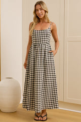 A woman modeling a black and white gingham maxi dress with spaghetti straps in a bright room with a large mirror, wooden floor, and cream-colored cabinets.