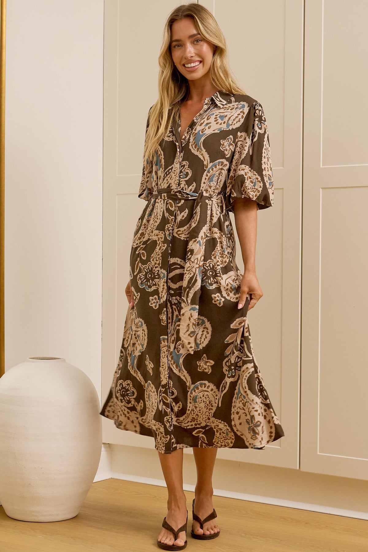 A woman wearing a brown and beige paisley midi dress stands in a bright room with cream closets and a large floor mirror, showing both her front and side profile. 