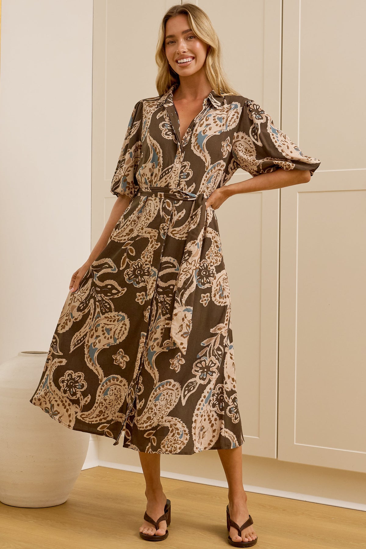 A woman models a brown and beige paisley midi dress in front of a white paneled wall and mirror, showing both the front and side profile of the dress. 