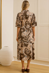 A woman stands with her back to the camera wearing a brown short-sleeved midi dress with a beige and blue paisley pattern, standing in front of white built-in wardrobes. A large floor-to-ceiling mirror on the left reflects her side profile.