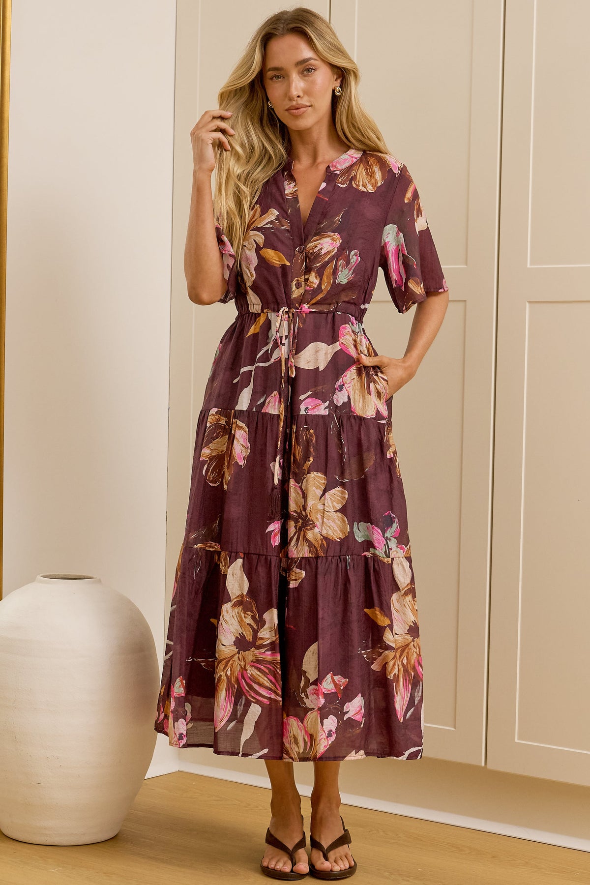 A woman stands in a bright room wearing a short-sleeve floral midi dress with a tie waist. She is positioned in front of cream-colored built-in wardrobes, with a large floor mirror on the left reflecting her side profile.