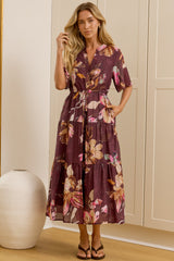 A woman stands in a bright room wearing a short-sleeve floral midi dress with a tie waist. She is positioned in front of cream-colored built-in wardrobes, with a large floor mirror on the left reflecting her side profile.