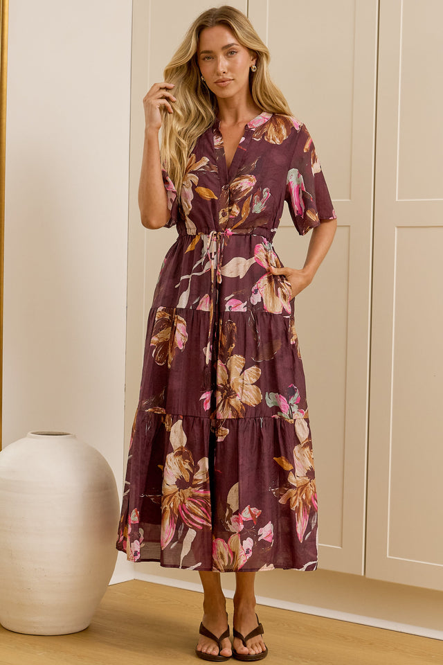 A woman stands in a bright room wearing a short-sleeve floral midi dress with a tie waist. She is positioned in front of cream-colored built-in wardrobes, with a large floor mirror on the left reflecting her side profile.