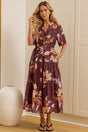 A woman stands in a bright room wearing a short-sleeve floral midi dress with a tie waist. She is positioned in front of cream-colored built-in wardrobes, with a large floor mirror on the left reflecting her side profile.