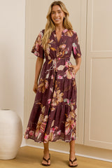 A woman wearing a short-sleeved, tiered midi dress with a floral print in purple, pink, and green stands in a room with cream-colored paneled walls and a large mirror showing her side profile.