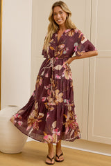A woman models a tiered, short-sleeved floral maxi dress in a dusty purple shade with pink and yellow floral patterns. She is standing in a bright room with cream-colored paneled walls, a large floor-to-ceiling mirror showing her side profile, and a white ceramic vase on a light wood floor.