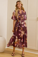 A woman models a purple floral midi wrap dress in a bright room with cream-colored wardrobes and a large mirror showing her profile. The dress features a purple base with pink, orange, and yellow floral patterns.