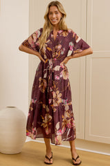 A woman modeling a short-sleeve, knee-length floral dress with a tie waist, standing in front of a large mirror and cream-colored wardrobe doors. The dress features a purple base with pink, orange, and yellow floral patterns.