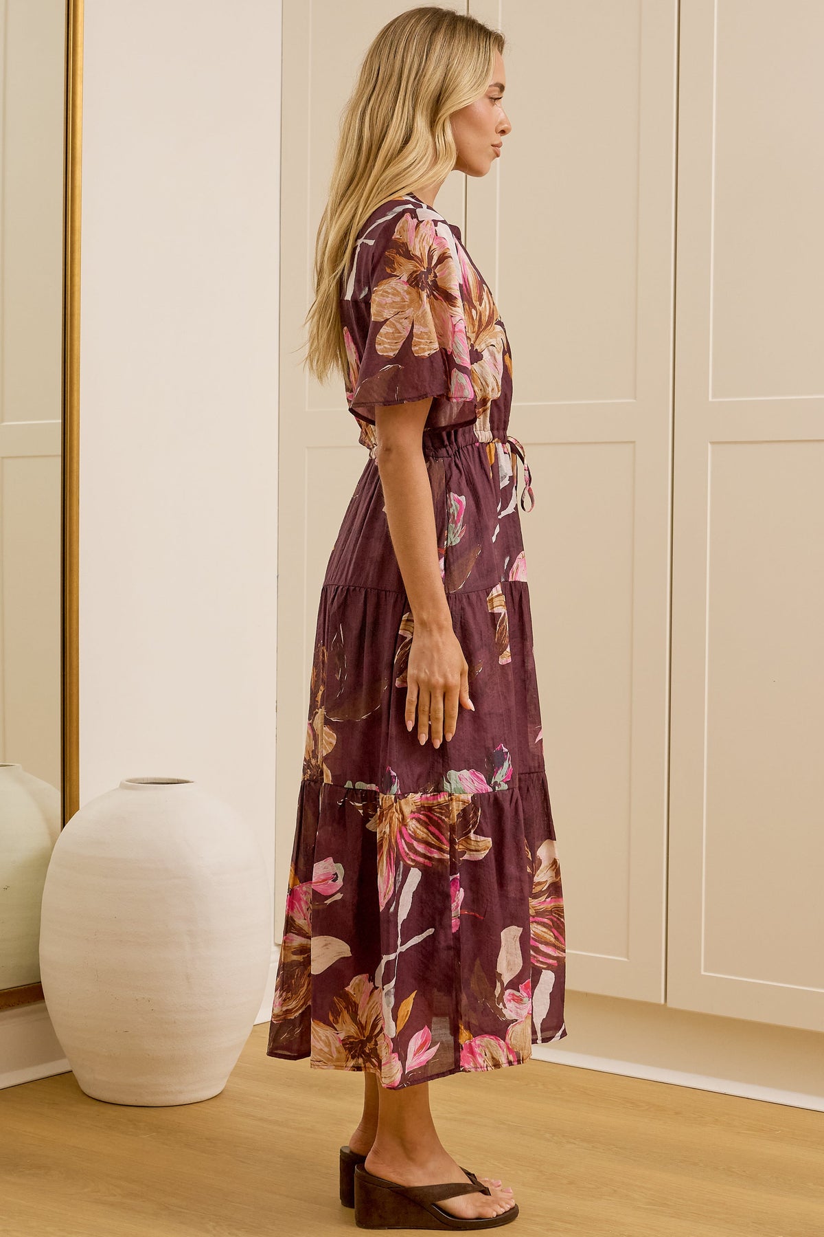 A woman stands in profile wearing a floral midi dress with short sleeves and a tie waist, standing in a bright room with cream-colored wardrobes and a large floor mirror reflecting her back.
