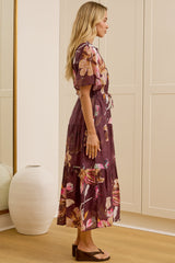 A woman stands in profile wearing a floral midi dress with short sleeves and a tie waist, standing in a bright room with cream-colored wardrobes and a large floor mirror reflecting her back.