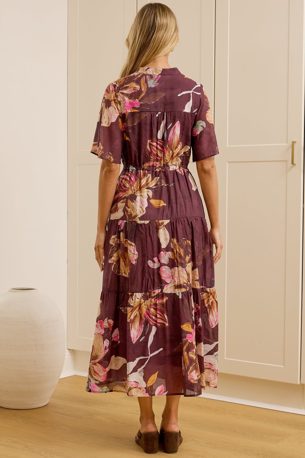 A woman stands with her back to the camera in a bright room, wearing a short-sleeved tiered maxi dress with a purple and pink floral print. She is positioned in front of white built-in wardrobes, with a large gold-framed mirror on the left reflecting her side profile.