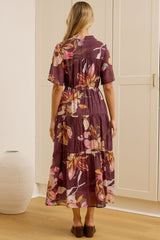 A woman stands with her back to the camera in a bright room, wearing a short-sleeved tiered maxi dress with a purple and pink floral print. She is positioned in front of white built-in wardrobes, with a large gold-framed mirror on the left reflecting her side profile.