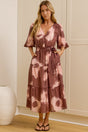 A woman stands in a bright room wearing a tiered midi dress with a dusty rose and pink tie-dye pattern. She is positioned in front of white paneled walls and a large floor-to-ceiling mirror, which reflects her side profile.