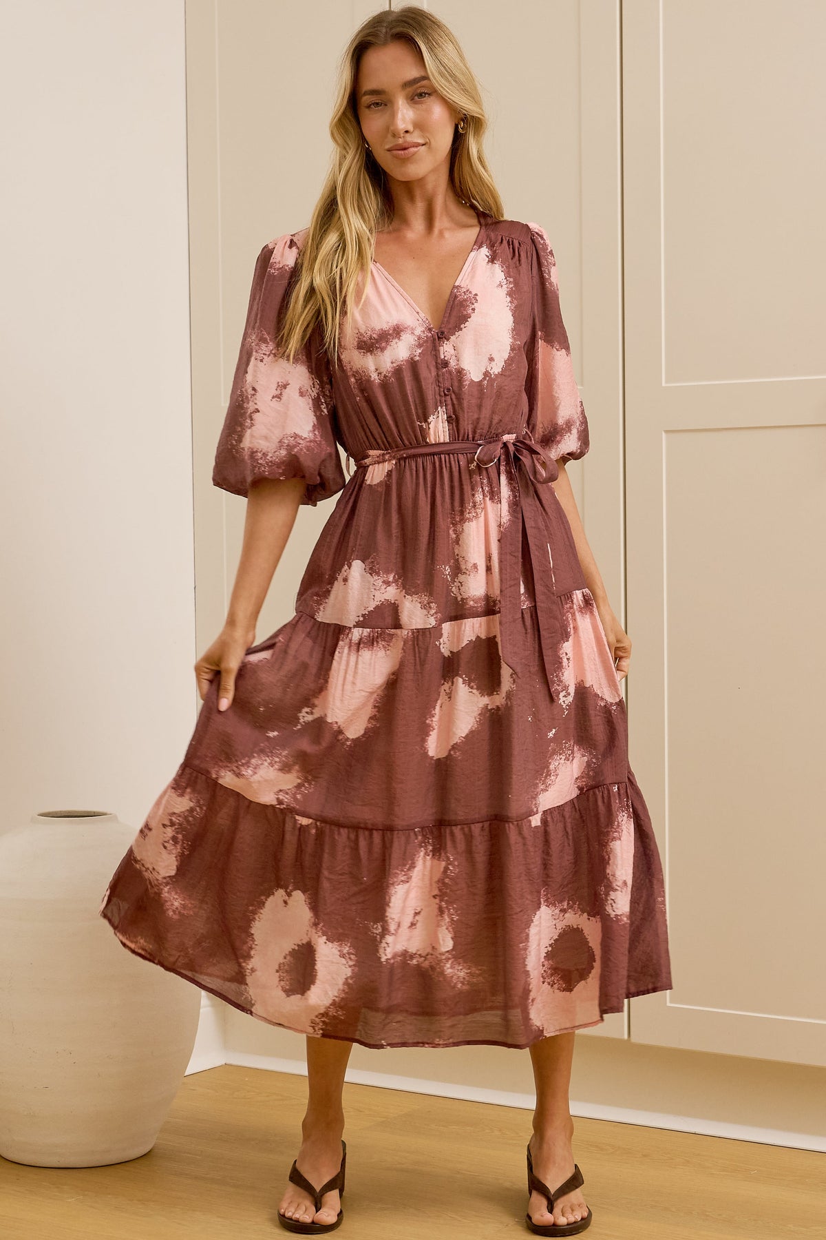 A woman models a tiered midi dress with a dusty rose and pink floral tie-dye pattern in a bright room with cream-colored cabinets and a large mirror showing her side profile.