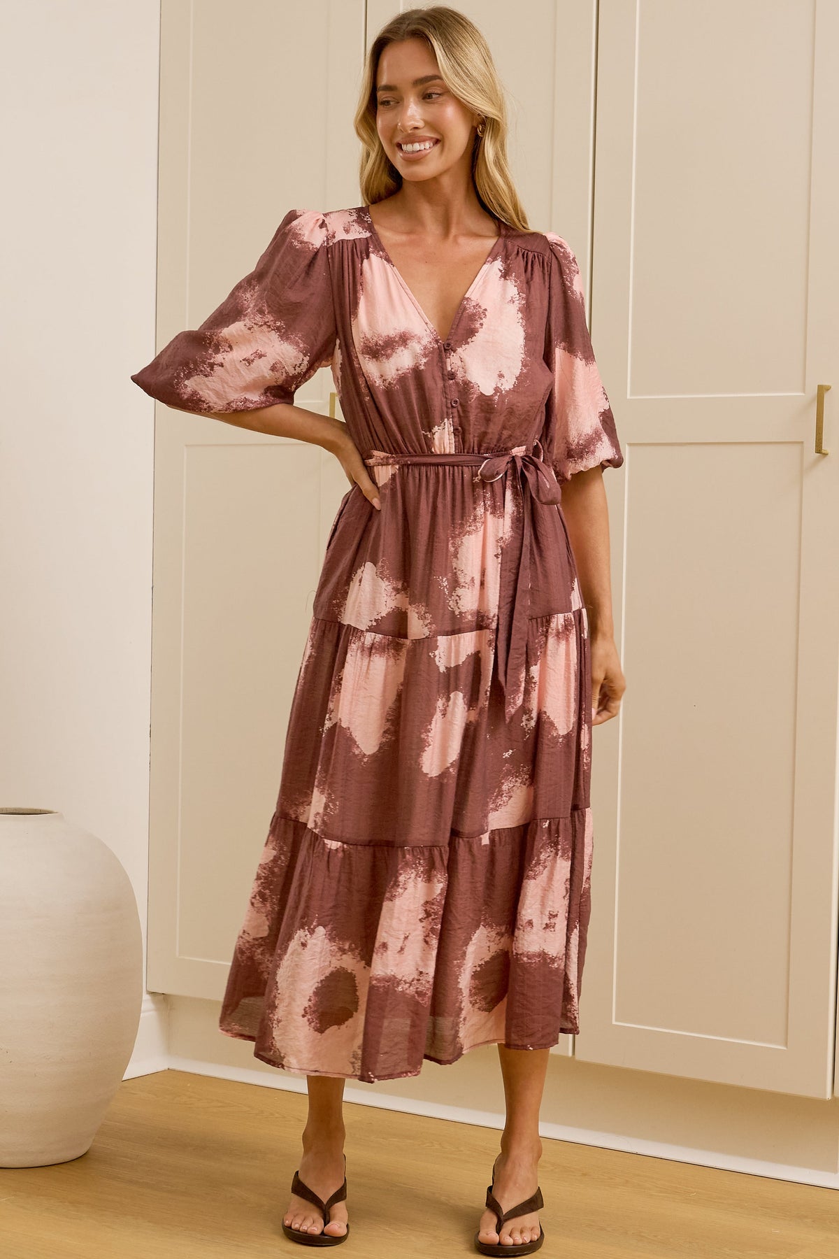 A woman modeling a tiered midi dress with a dusty rose and pink abstract floral pattern, standing in a room with light wood floors, white paneled walls, and a large mirror showing her side profile. 