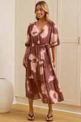 A woman models a tiered midi dress with a dusty rose and pink tie-dye pattern in a bright room with white wardrobes and a large mirror showing her reflection.