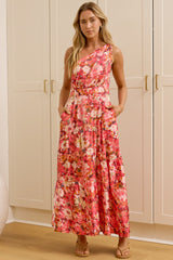 A woman stands in a bright room wearing a sleeveless, floor-length floral maxi dress. A large floor mirror on the left reflects her side profile, and white paneled walls and doors are behind her.