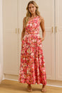 A woman stands in a bright room wearing a sleeveless, floor-length floral maxi dress. A large floor mirror on the left reflects her side profile, and white paneled walls and doors are behind her.