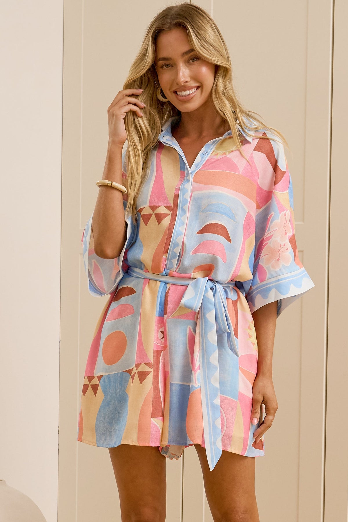 A woman stands in a bedroom wearing a pastel multicolor print robe dress with a tie waist and elbow-length sleeves. She is wearing beige slide sandals, with a large floor mirror on the left reflecting her profile.