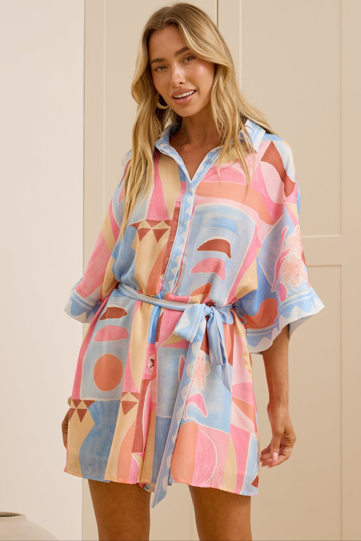 A woman modeling a short, button-front shirtdress with a tie waist, featuring a colorful abstract geometric print in pink, blue, and beige, set against a background of cream-colored paneled doors.