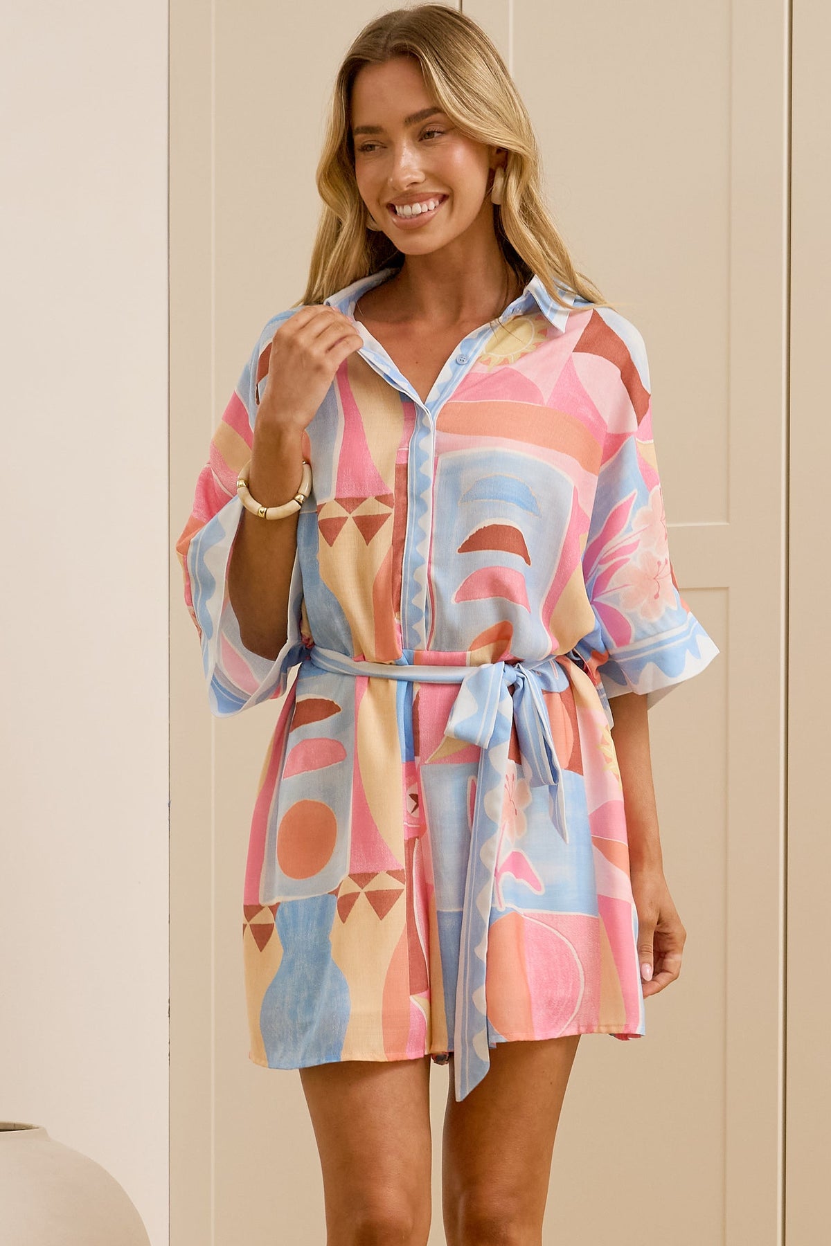 A woman stands in a bright room wearing a short, colorful abstract-patterned shirt dress with a tie waist. She is wearing beige slide sandals, and a large mirror on the left reflects her side profile.