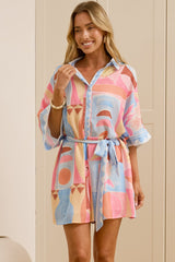 A woman stands in a bright room wearing a short, colorful abstract-patterned shirt dress with a tie waist. She is wearing beige slide sandals, and a large mirror on the left reflects her side profile.