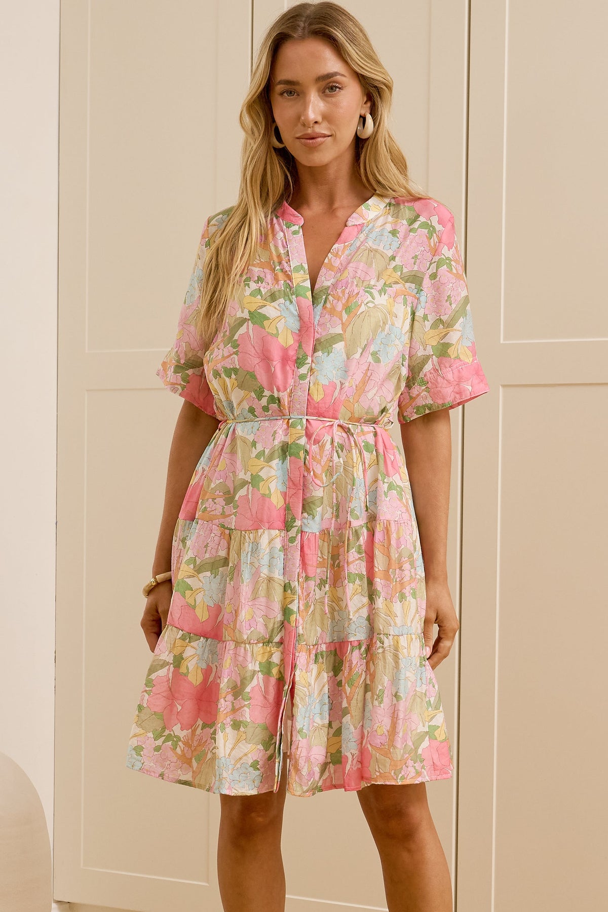 A woman modeling a short-sleeve pink floral dress in a bright room with cream-colored cabinets and a large mirror showing her side profile.