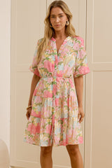 A woman modeling a short-sleeve pink floral dress in a bright room with cream-colored cabinets and a large mirror showing her side profile.