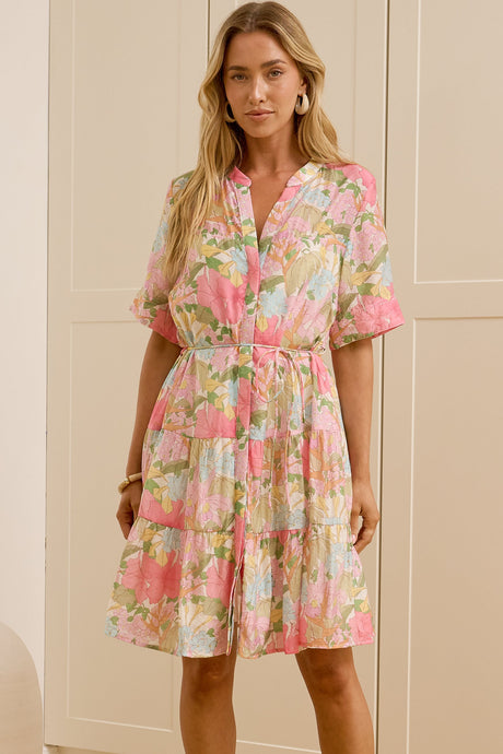 A woman modeling a short-sleeve pink floral dress in a bright room with cream-colored cabinets and a large mirror showing her side profile.