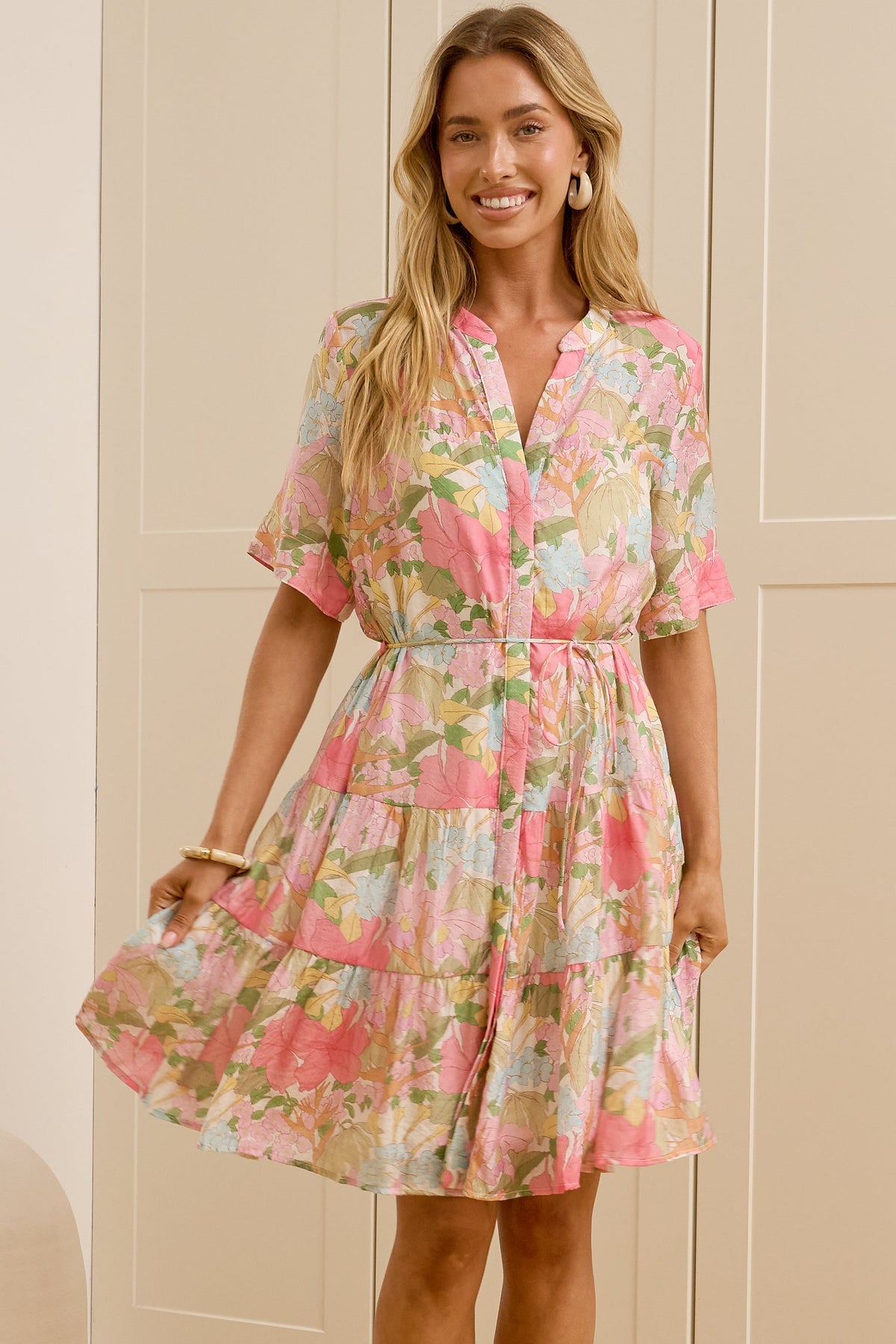 A woman stands in a bright room wearing a short-sleeve floral dress with a pink, green, and yellow pattern. She is standing in front of cream-colored wardrobe doors, with her reflection visible in a mirror on the left.