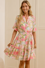A woman stands in a bright room wearing a short-sleeve floral dress with a pink, green, and yellow pattern. She is standing in front of cream-colored wardrobe doors, with her reflection visible in a mirror on the left.