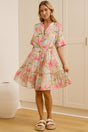 A woman stands in a bright room wearing a short-sleeve tiered floral dress with pink, green, and beige tones. A large mirror on the left reflects her side profile, and cream-colored cabinets line the background wall.